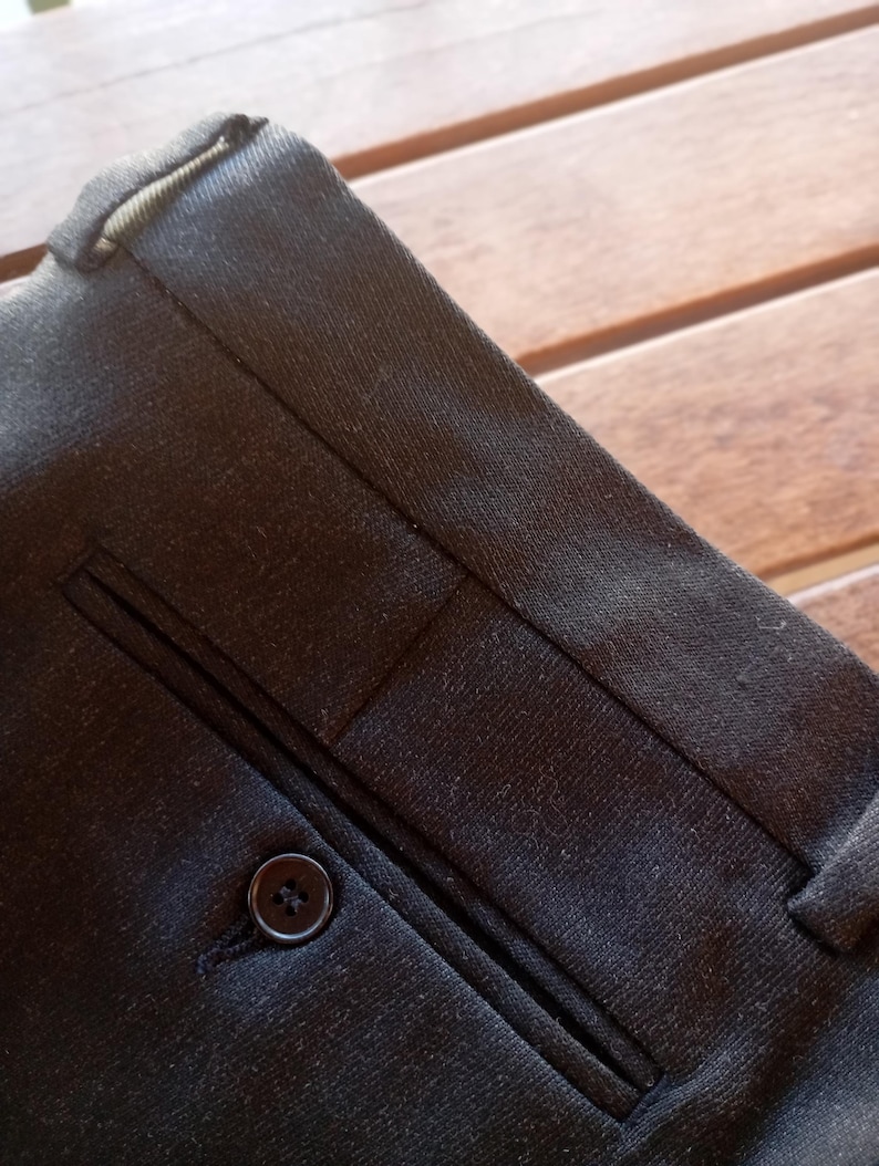 May include: A pair of dark charcoal grey trousers featuring a structured waistband with belt loops and a single welt pocket on the front. The pocket is secured with a small, round, dark-coloured button centred on the horizontal opening.