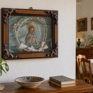 May include: An antique framed religious icon featuring a silver relief of a Madonna and child, set against a teal background. The ornate, dark wood frame is mounted on a white wall. A terracotta pot with a green plant, a decorative bowl, and books are on a wooden table.