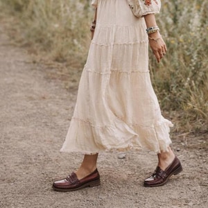 May include: A cream-coloured tiered maxi skirt paired with burgundy leather loafers. The skirt has a flowing, ruffled design. The loafers have a classic penny loafer style with a low heel, suitable for a casual or bohemian aesthetic.