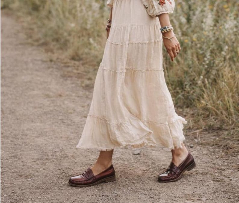 May include: A cream-coloured tiered maxi skirt paired with burgundy leather loafers. The skirt has a flowing, ruffled design. The loafers have a classic penny loafer style with a low heel, suitable for a casual or bohemian aesthetic.