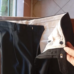 May include: A pair of black trousers featuring a high waistband with a button closure and a metal hook-and-eye fastener. The interior of the waistband is lined with a white fabric featuring a subtle pinstripe pattern and a dark button.