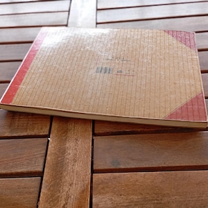 Op de afbeelding: Een vintage notitieboek met een bruine kaft en rode rug- en hoekaccenten. Het notitieboek ligt op een houten tafel met een lattenontwerp. De kaft heeft een getextureerde, gestreepte uitstraling.