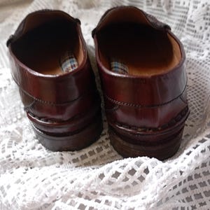 May include: A pair of brown leather loafers. The shoes have a polished finish and a stacked heel. The interior features a plaid pattern. The shoes are displayed on a white, open-weave fabric. The shoes are a classic design.