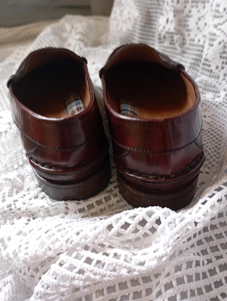 May include: A pair of brown leather loafers. The shoes have a polished finish and a stacked heel. The interior features a plaid pattern. The shoes are displayed on a white, open-weave fabric. The shoes are a classic design.