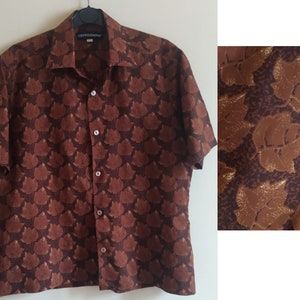 Puede incluir: Una camisa de manga corta de color marr&oacute;n con botones y un estampado de hojas doradas. La camisa tiene cuello y cierre de botones.