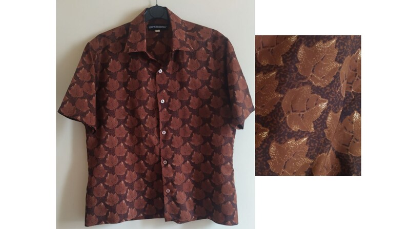 Puede incluir: Una camisa de manga corta de color marr&oacute;n con botones y un estampado de hojas doradas. La camisa tiene cuello y cierre de botones.