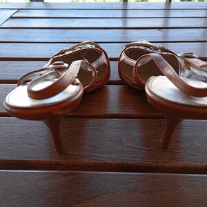May include: A pair of brown leather heeled sandals with a white strap and buckle detail. The sandals are on a wooden surface.
