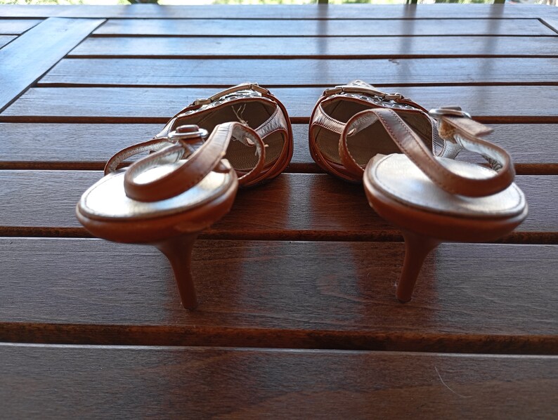 May include: A pair of brown leather heeled sandals with a white strap and buckle detail. The sandals are on a wooden surface.
