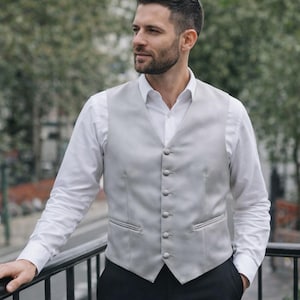 May include: A light grey waistcoat with matching buttons, worn over a white shirt. The waistcoat has two front pockets and is paired with black trousers. The person is standing on a balcony with a black railing.