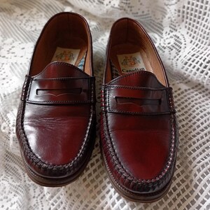 May include: A pair of burgundy leather penny loafers. The shoes feature a classic design with a stitched sole and a strap across the vamp. The word "Canadian" is printed inside. The shoes are displayed on a white lace tablecloth.