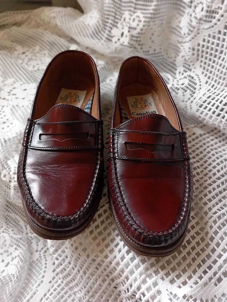 May include: A pair of burgundy leather penny loafers. The shoes feature a classic design with a stitched sole and a strap across the vamp. The word "Canadian" is printed inside. The shoes are displayed on a white lace tablecloth.