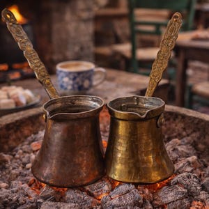 May include: Two copper and brass coffee pots, or cezves, sit on hot embers. The pots have ornate handles and are set in a stone hearth. A cup of coffee and a plate of sweets are in the background, suggesting a cafe setting.