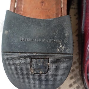 May include: A close-up of a shoe sole with the text "IMPORT-PLAST HELLAS" and a logo. The sole is black with a textured pattern. The shoe's upper is a burgundy colour.