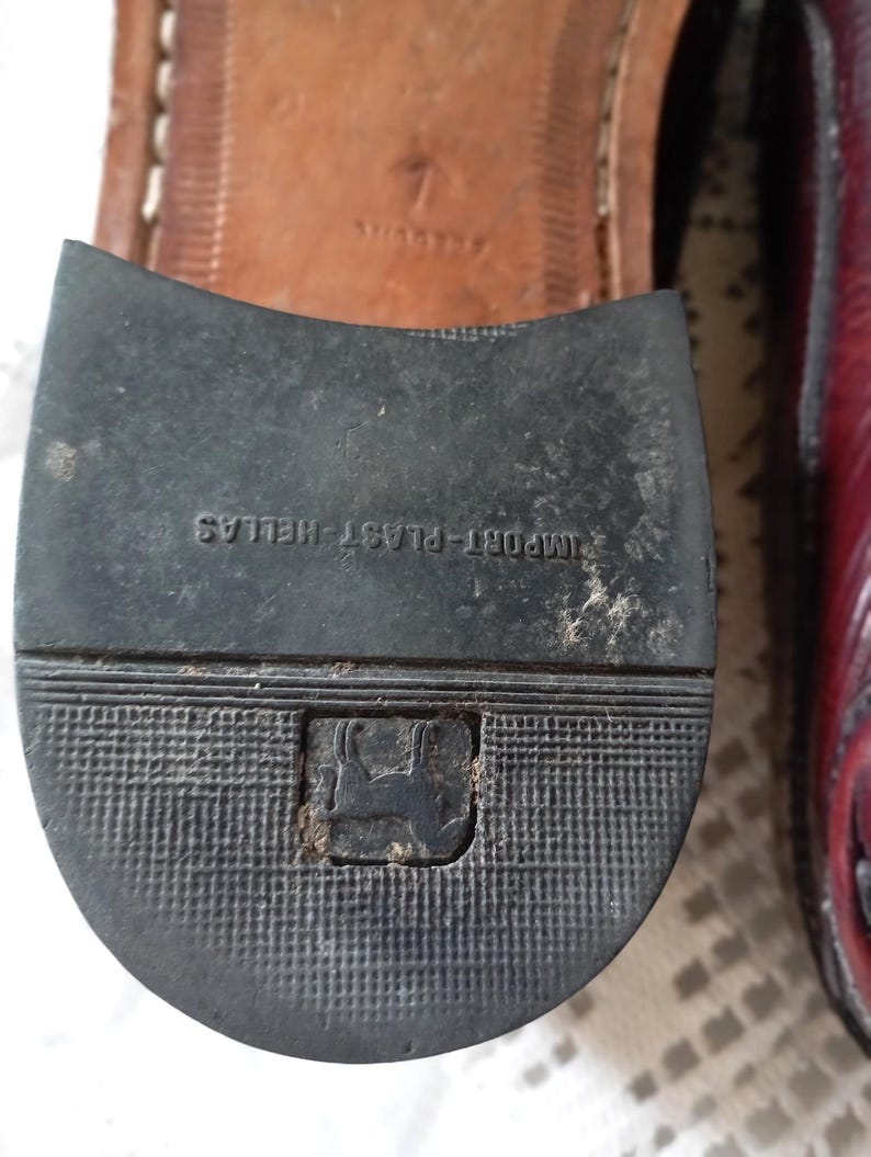May include: A close-up of a shoe sole with the text "IMPORT-PLAST HELLAS" and a logo. The sole is black with a textured pattern. The shoe's upper is a burgundy colour.