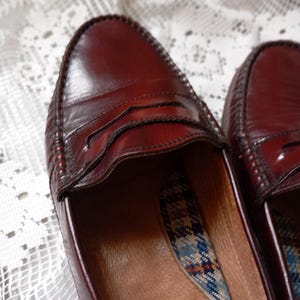 May include: A pair of burgundy leather penny loafers. The shoes have a classic design with a stitched vamp and a decorative strap across the top. The interior features a plaid pattern and a label that says "Canadian".