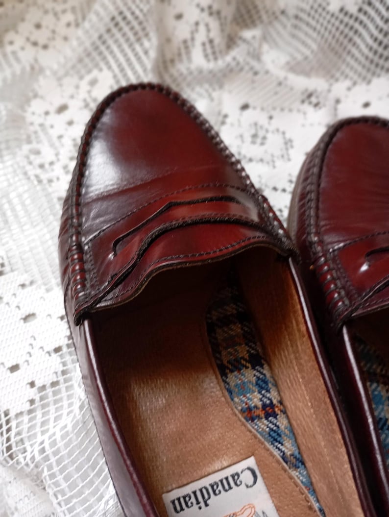 May include: A pair of burgundy leather penny loafers. The shoes have a classic design with a stitched vamp and a decorative strap across the top. The interior features a plaid pattern and a label that says "Canadian".