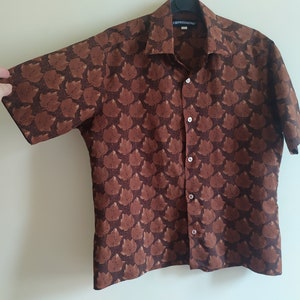 Puede incluir: Una camisa marr&oacute;n de manga corta con botones y un patr&oacute;n repetido de caras estilizadas marrones. La camisa tiene cuello y cierre de botones. La etiqueta de la camisa dice "EXPRESSIONS".