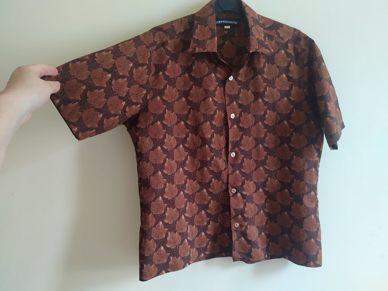 Puede incluir: Una camisa marr&oacute;n de manga corta con botones y un patr&oacute;n repetido de caras estilizadas marrones. La camisa tiene cuello y cierre de botones. La etiqueta de la camisa dice "EXPRESSIONS".