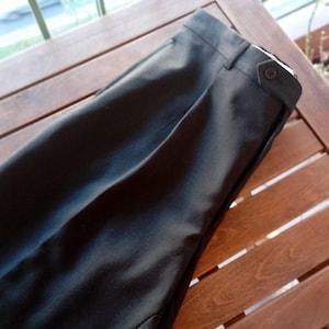 May include: A pair of black trousers featuring a structured waistband with a side tab adjustment. The trousers include a single button closure on the side tab, belt loops, and a clean, pressed front crease running down the length of the leg fabric.