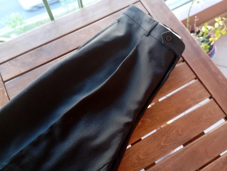May include: A pair of black trousers featuring a structured waistband with a side tab adjustment. The trousers include a single button closure on the side tab, belt loops, and a clean, pressed front crease running down the length of the leg fabric.