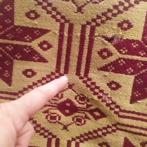 May include: Close-up of a red and gold woven fabric with a tear in the centre. The fabric has a geometric pattern.