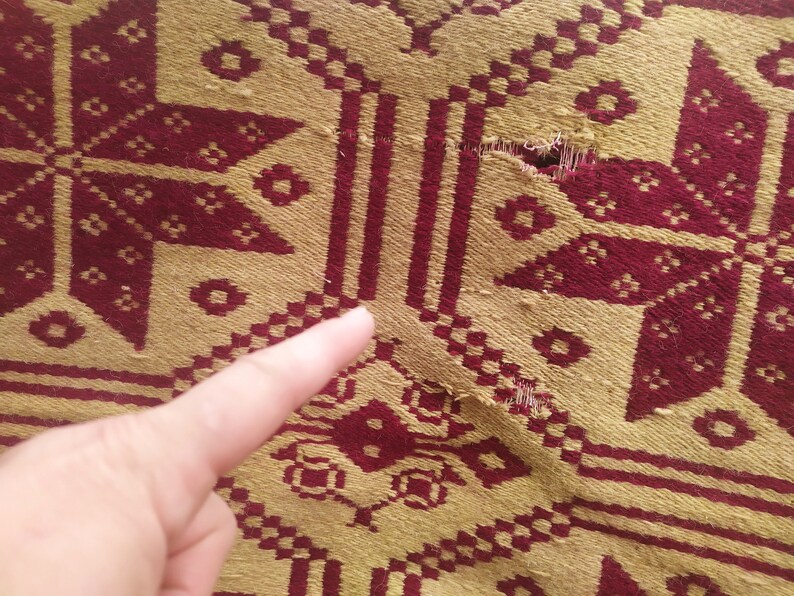 May include: Close-up of a red and gold woven fabric with a tear in the centre. The fabric has a geometric pattern.