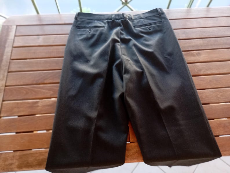 May include: A pair of black trousers featuring a waistband with belt loops, a centre front zip closure, and two back pockets with button closures. The trousers have a straight-leg cut with a visible crease running down the centre of each leg.
