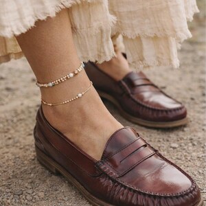 May include: Brown leather loafers with a penny strap, worn with a layered gold anklet. The anklet features small beads and pearls. The shoes are on a gravel surface. The loafers are a classic style.