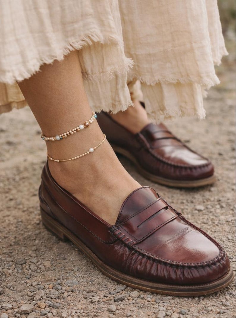 May include: Brown leather loafers with a penny strap, worn with a layered gold anklet. The anklet features small beads and pearls. The shoes are on a gravel surface. The loafers are a classic style.