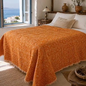Pode incluir: Uma colcha laranja vibrante com um padrão geométrico e floral repetitivo e um delicado acabamento de franjas nas bordas. O tecido está estendido sobre uma cama, exibindo a sua superfície texturizada e o design intrincado num tom quente.