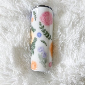 Op de afbeelding: Een witte roestvrijstalen beker met een bloemenmotief. Het ontwerp bevat roze, gele, blauwe en groene bloemen en bladeren op een witte achtergrond.