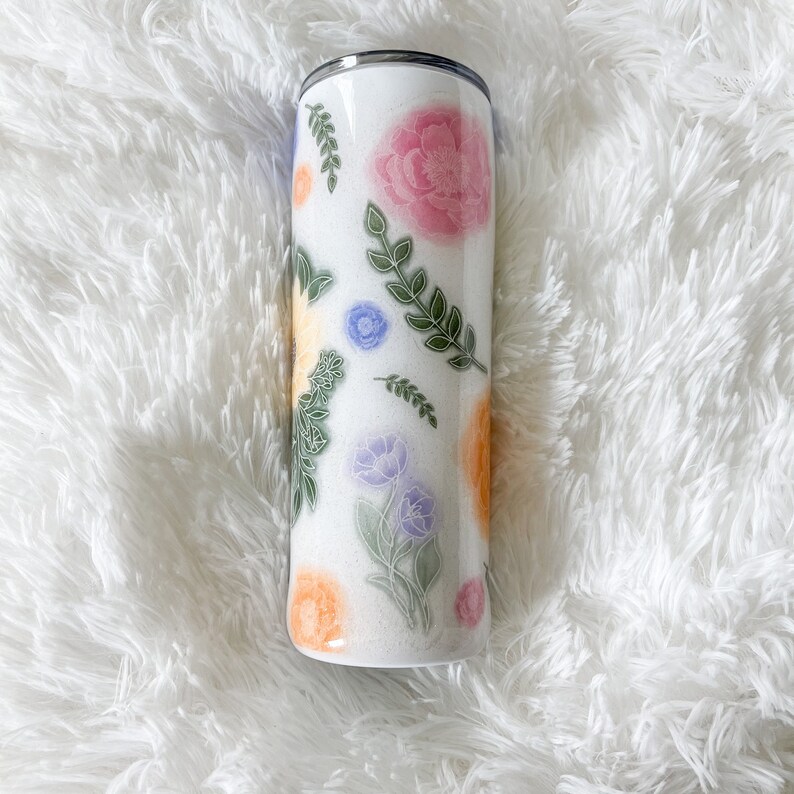 Op de afbeelding: Een witte roestvrijstalen beker met een bloemenmotief. Het ontwerp bevat roze, gele, blauwe en groene bloemen en bladeren op een witte achtergrond.
