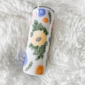 Op de afbeelding: Een witte roestvrijstalen beker met een bloemenpatroon. Het patroon bevat gele, oranje, blauwe en roze bloemen met groene bladeren.