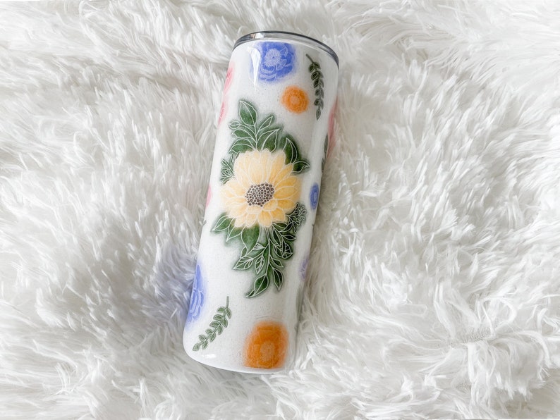 Op de afbeelding: Een witte roestvrijstalen beker met een bloemenpatroon. Het patroon bevat gele, oranje, blauwe en roze bloemen met groene bladeren.