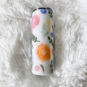 Op de afbeelding: Een witte roestvrijstalen beker met een bloemenontwerp met roze, oranje, gele, blauwe en groene bloemen en bladeren.