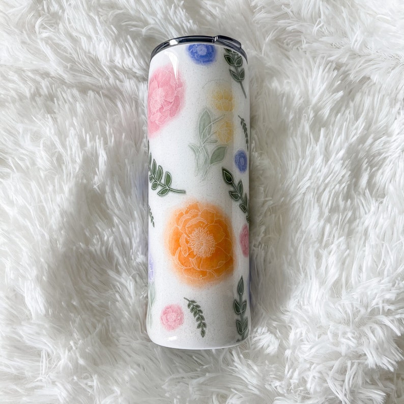 Op de afbeelding: Een witte roestvrijstalen beker met een bloemenontwerp met roze, oranje, gele, blauwe en groene bloemen en bladeren.