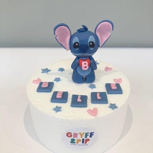 Peut inclure: Un cake topper repr&eacute;sentant un personnage de dessin anim&eacute; bleu tenant un c&oelig;ur avec la lettre "B". Le g&acirc;teau est d&eacute;cor&eacute; de carr&eacute;s bleus &eacute;pelant "BELLE", de c&oelig;urs roses et d'&eacute;toiles bleues. La base porte le logo "GRYFF & PIP CAKE TOPPERS".