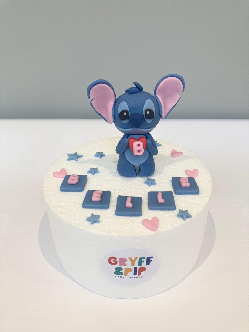 Peut inclure: Un cake topper repr&eacute;sentant un personnage de dessin anim&eacute; bleu tenant un c&oelig;ur avec la lettre "B". Le g&acirc;teau est d&eacute;cor&eacute; de carr&eacute;s bleus &eacute;pelant "BELLE", de c&oelig;urs roses et d'&eacute;toiles bleues. La base porte le logo "GRYFF & PIP CAKE TOPPERS".