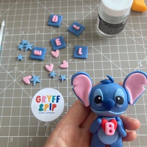 Peut inclure: Un cake topper Stitch bleu avec des oreilles roses et un c&oelig;ur rouge avec la lettre "B". Des carr&eacute;s bleus avec les lettres roses "BEL", des c&oelig;urs roses et des &eacute;toiles bleues sont &eacute;parpill&eacute;s. Un autocollant "GRYFF & PIP CAKE TOPPERS" est visible.
