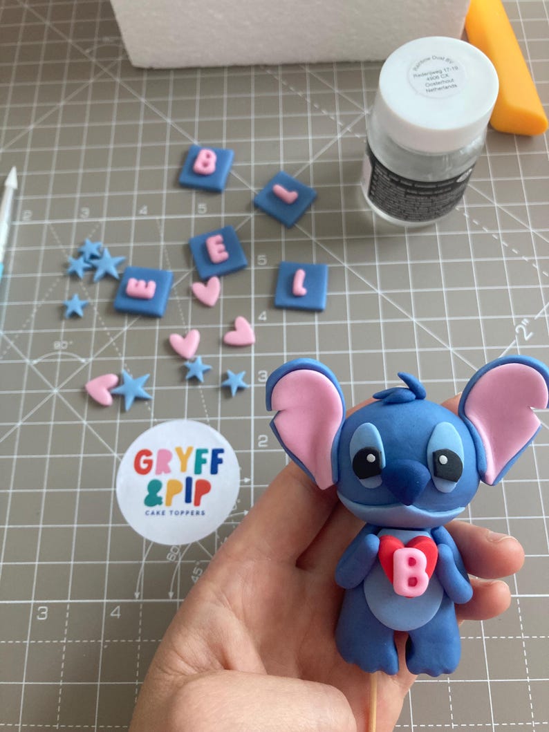Peut inclure: Un cake topper Stitch bleu avec des oreilles roses et un c&oelig;ur rouge avec la lettre "B". Des carr&eacute;s bleus avec les lettres roses "BEL", des c&oelig;urs roses et des &eacute;toiles bleues sont &eacute;parpill&eacute;s. Un autocollant "GRYFF & PIP CAKE TOPPERS" est visible.