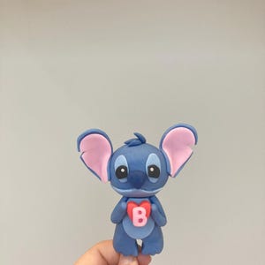 Peut inclure: Figurine bleue repr&eacute;sentant un personnage de dessin anim&eacute; avec de grandes oreilles et un embl&egrave;me en forme de c&oelig;ur sur la poitrine. L'embl&egrave;me porte la lettre "B" en rose. Le personnage a de grands yeux et tient le c&oelig;ur.