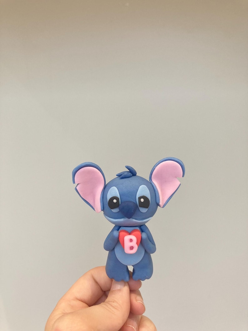 Peut inclure: Figurine bleue repr&eacute;sentant un personnage de dessin anim&eacute; avec de grandes oreilles et un embl&egrave;me en forme de c&oelig;ur sur la poitrine. L'embl&egrave;me porte la lettre "B" en rose. Le personnage a de grands yeux et tient le c&oelig;ur.