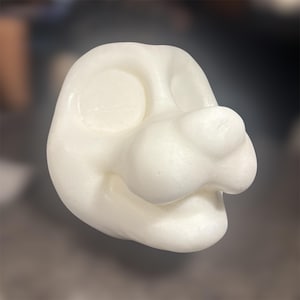 Puede incluir: Una base de cabeza de animal de espuma blanca moldeada con un hocico redondeado, una nariz prominente y dos cuencas oculares circulares hundidas. La superficie tiene un aspecto texturizado y poroso propio de la espuma de poliestireno.