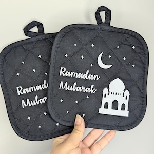 May include: Two black quilted pot holders with white text that reads "Ramadan Mubarak". The pot holders feature a white crescent moon, stars, and a mosque design. Each pot holder has a small black loop for hanging.