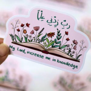 May include: A pink sticker with a floral design and text in English and Arabic. The sticker features an open book with colorful flowers and the phrase "My Lord, increase me in knowledge." The Arabic text is in green.