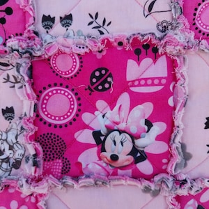 Puede incluir: Primer plano de un edred&oacute;n de retazos rosa y blanco con un tema de Minnie Mouse. El edred&oacute;n presenta cuadrados con Minnie Mouse, flores y lunares. Los bordes tienen un estilo deshilachado, de edred&oacute;n de trapo.
