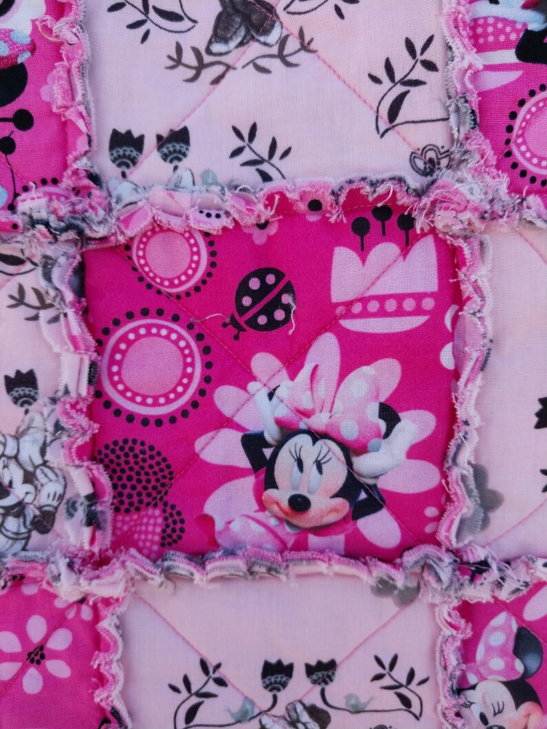Puede incluir: Primer plano de un edred&oacute;n de retazos rosa y blanco con un tema de Minnie Mouse. El edred&oacute;n presenta cuadrados con Minnie Mouse, flores y lunares. Los bordes tienen un estilo deshilachado, de edred&oacute;n de trapo.