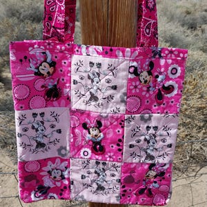 Puede incluir: Un bolso tote acolchado rosa con un dise&ntilde;o de patchwork con ilustraciones de Minnie Mouse. El bolso tiene asas a juego con estampado de cachemira rosa. El bolso es de tela y tiene forma rectangular.