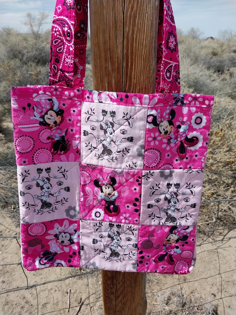 Puede incluir: Un bolso tote acolchado rosa con un dise&ntilde;o de patchwork con ilustraciones de Minnie Mouse. El bolso tiene asas a juego con estampado de cachemira rosa. El bolso es de tela y tiene forma rectangular.