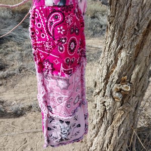 Puede incluir: Un bolso tote rosa hecho a mano con un dise&ntilde;o de patchwork. El bolso presenta varios patrones de cachemira y florales en tonos de rosa, blanco y negro. El bolso est&aacute; colgado de una rama de &aacute;rbol.